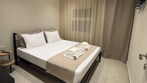 Una cama bien tendida con una colcha color beige y almohadas, una mesita de noche con un control remoto y una ventana con cortinas traslúcidas.