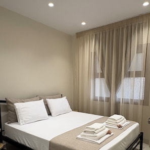 Una cama bien hecha con sábanas y almohadas blancas, una colcha color beige y una pila de toallas dobladas en el cabecero.