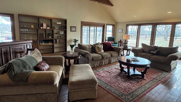 A spacious living room with multiple seating areas, a wooden ceiling, large windows, and a bookshelf filled with books and decorative items. - Large private home in gated horse farm in the heart of horse country! (Lexington)