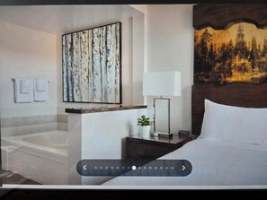 Een hotelkamer met een groot bed, een schilderij van bomen, een nachtkastje met een lamp en een potplant.