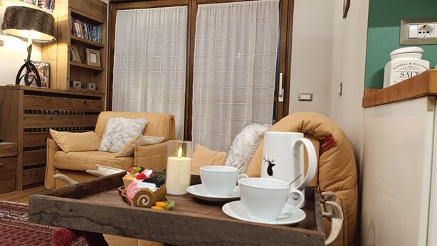 A cozy living room with a wooden sofa, a small table with cups and a candle, and a bookshelf filled with books.