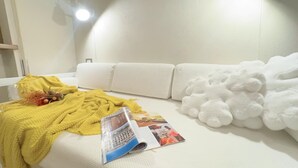 A white sofa with a yellow blanket and a magazine, a white wall, and a white fluffy object on the right.