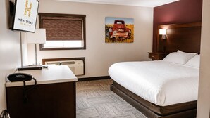 A hotel room with a bed, a desk with a telephone, a radiator, a window with blinds, and a wall-mounted artwork. - Homestead Inn and Suites (Vanderhoof)