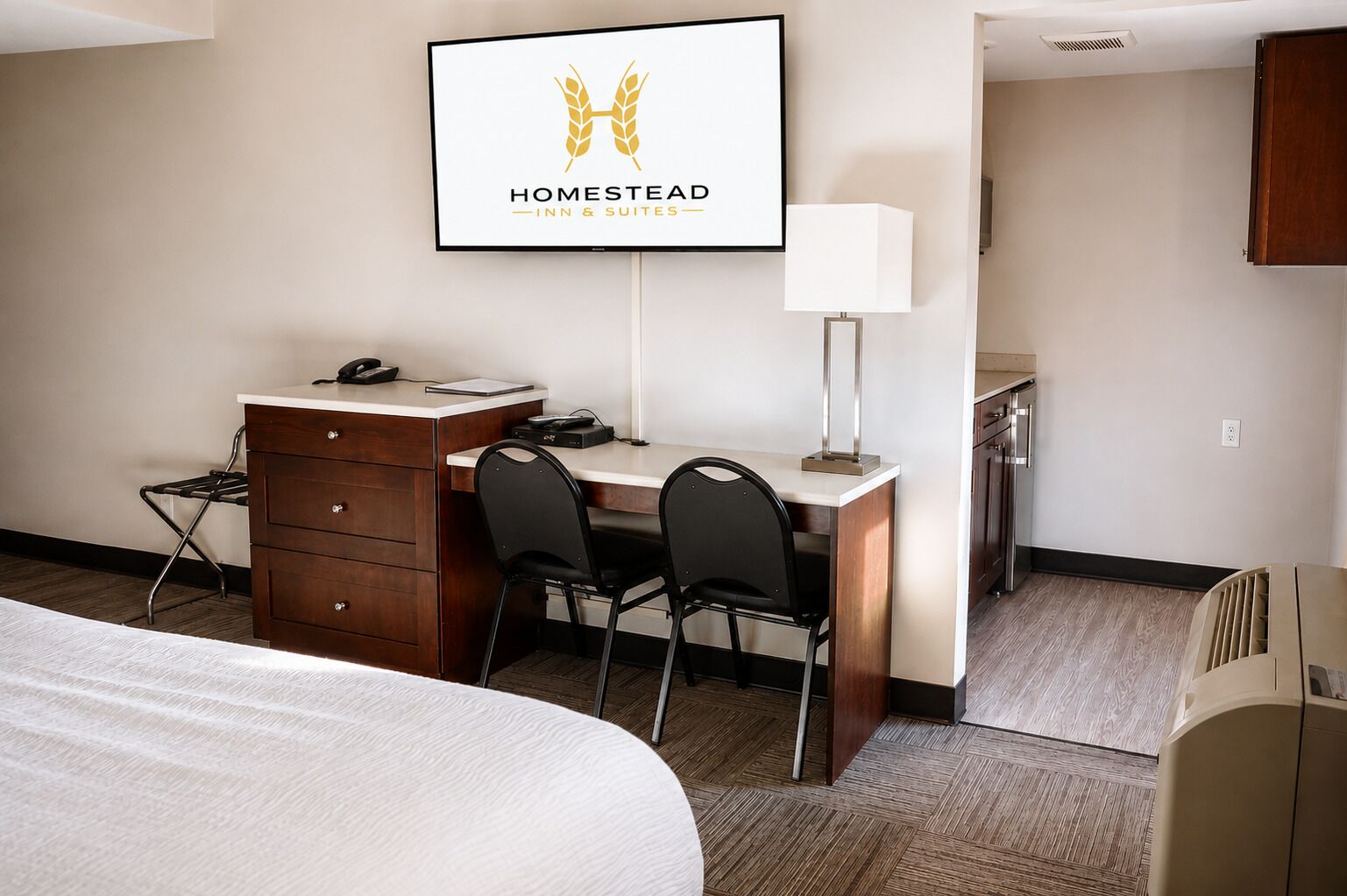 A hotel room with a bed, a desk with a chair, a telephone, a lamp, and a framed sign on the wall.