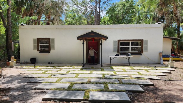 Un edificio pequeño de una planta con puerta roja, ventana con contraventanas y una pequeña mesa con una silla afuera.