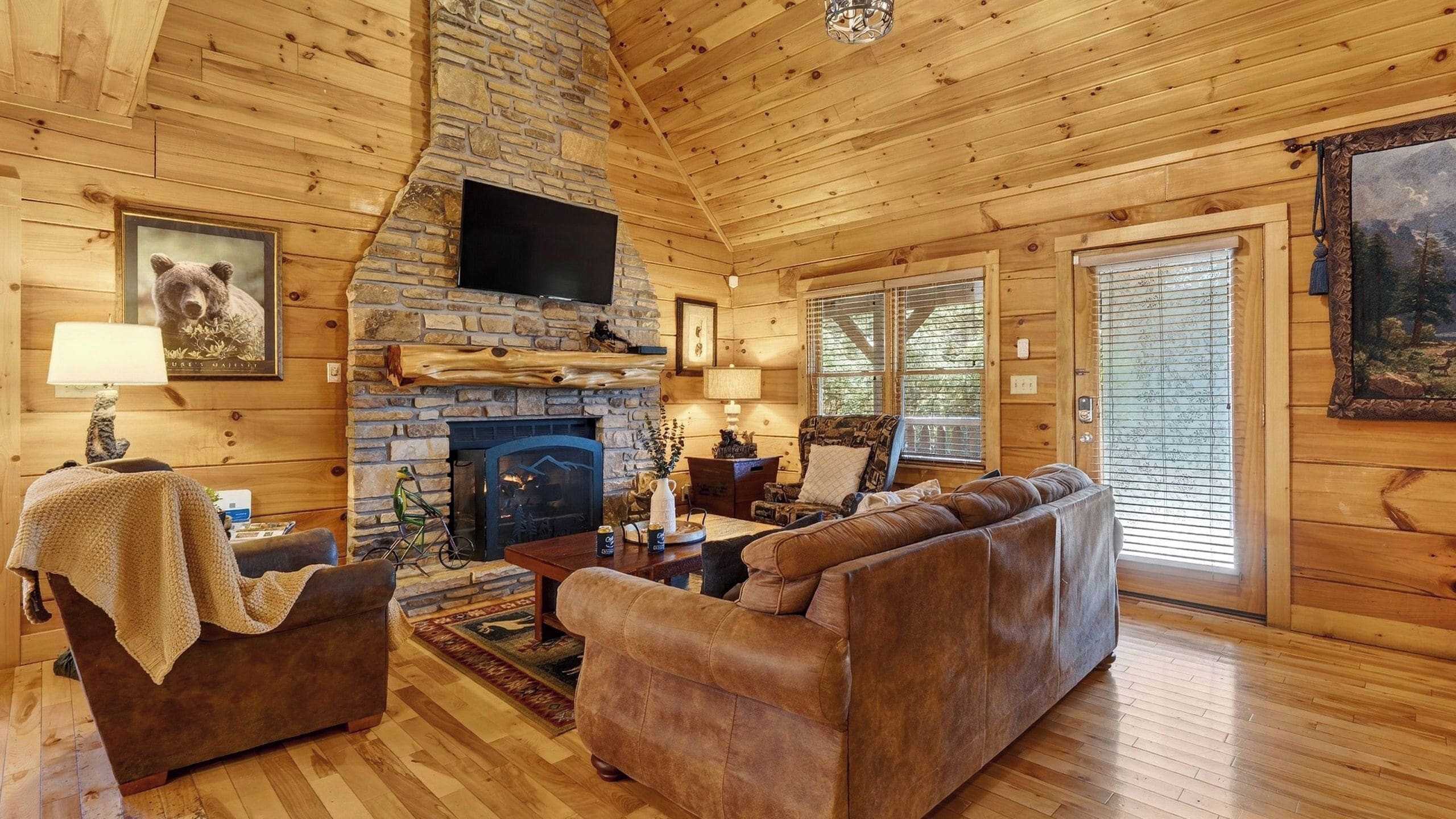 A cozy log cabin interior with a stone fireplace, wooden walls, a television mounted above it, and a large window.