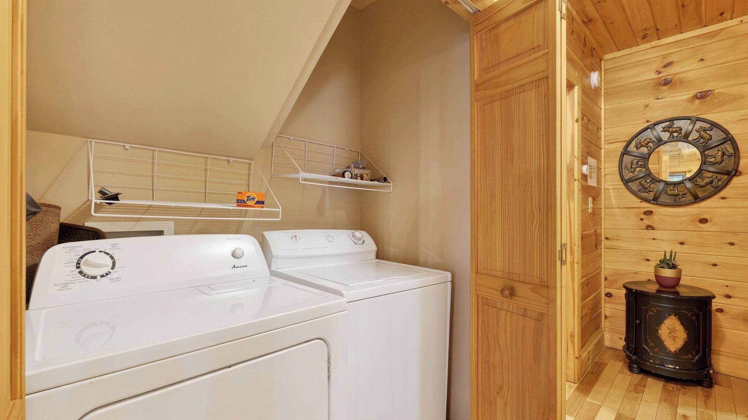 A laundry area with two white front-loading washers and dryers, a shelf with laundry supplies, and a rustic wooden wall with a decorative mirror and a small table with a plant.