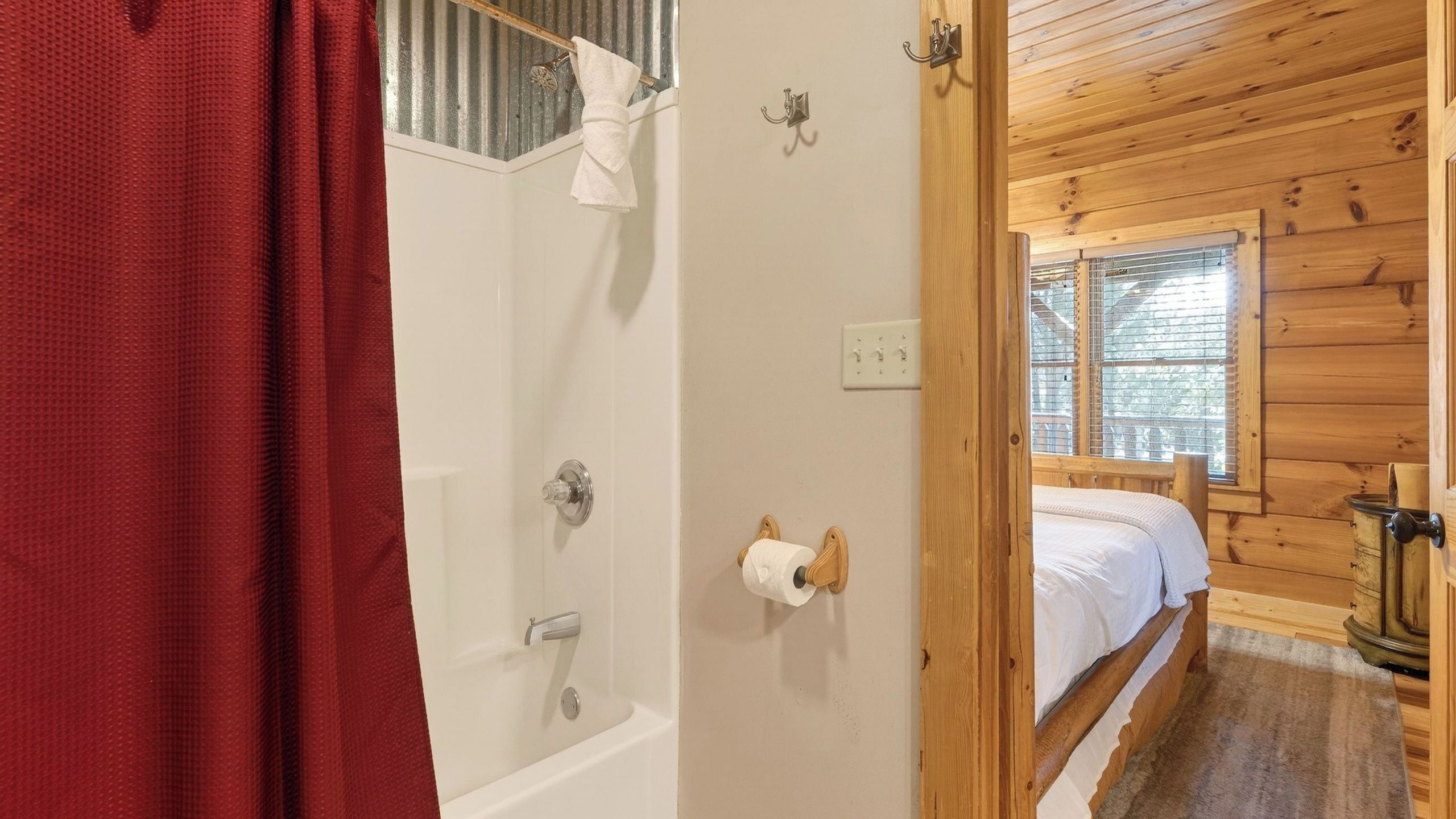 Ein Schlafzimmer in einer Holzhütte mit einem hölzernen Bett, einem Fenster mit Jalousien sowie einem Badezimmer mit Dusche, Toilette und Handtuchhalter.