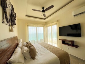 Habitación de hotel con cama, ventilador de techo, televisión y vista al océano.