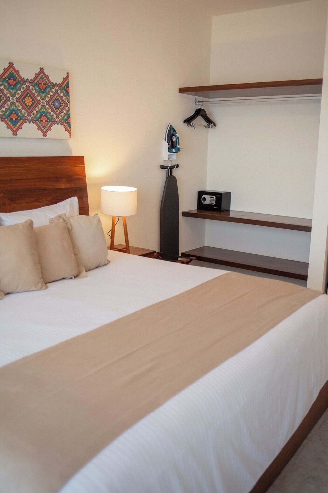 Un lit bien fait, recouvert d’une couvre-lit beige, avec une tête de lit en bois et une étagère fixée au mur, sur laquelle se trouvent une radio et un fer à repasser.