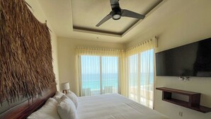 Ein Hotelzimmer mit Bett, Deckenventilator, Fernseher, einem Fenster mit Vorhängen und Meerblick.