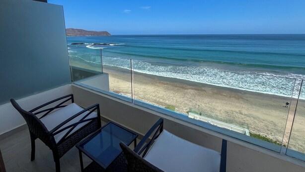 Ein Balkon mit Glastisch und Stühlen, der Blick auf einen Strand mit Wellen und sanften Hügeln im Hintergrund bietet.