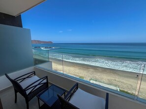 Un balcon avec une table et des chaises en verre, donnant sur une plage où l’on peut voir les vagues et des collines au loin.