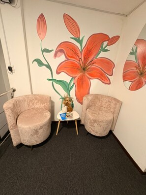 Um quarto com duas poltronas bege, uma pequena mesa branca com um vaso e livros, e uma parede com grandes murais de flores laranja.