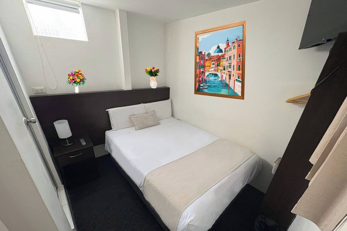 A hotel room with a bed, bedside tables, a painting, and a window.