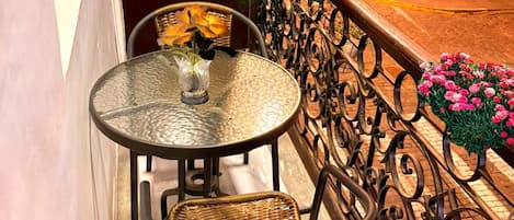 A small outdoor balcony with a round glass table, two wicker chairs, and a potted plant with pink flowers.