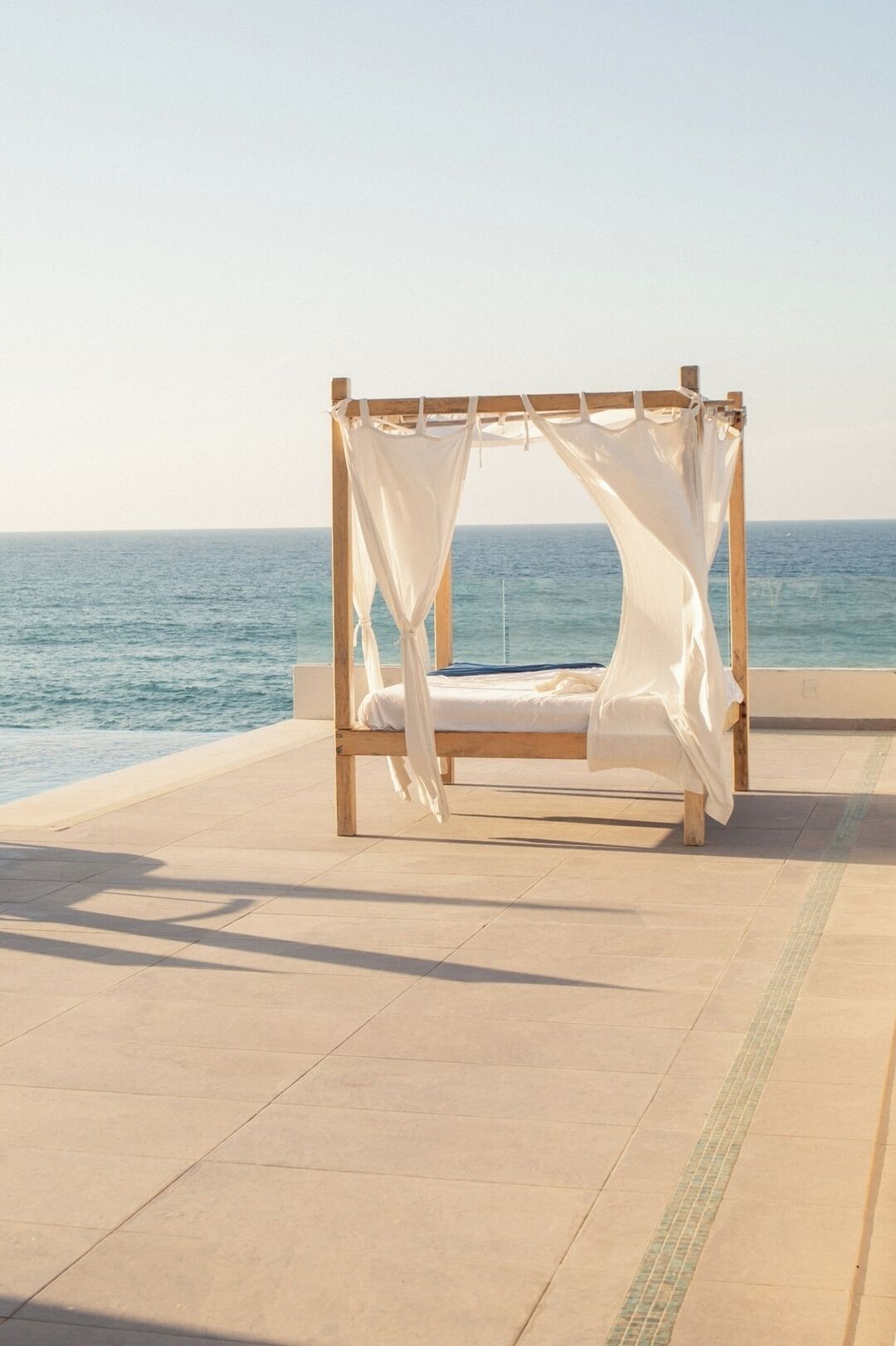 Estructura de cama de madera con ropa de cama blanca, sobre piso de baldosas y vista al mar.