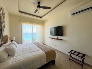 Una camera d'albergo con un letto, una ventola a soffitto, una TV a muro e vista sull'oceano.