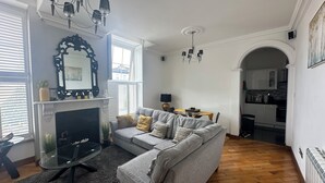 Una sala de estar con un sofá gris, televisor y chimenea. Hay un comedor con mesa y sillas, y una cocina visible a través de un arco.