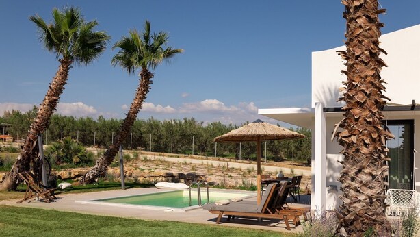 A modern outdoor pool area with lounge chairs and a thatched umbrella, surrounded by palm trees and a white building. - Exotic Villa, Private Pool with Lagoon Views – Romantic Escape in Halkidiki (Nea Moudania)