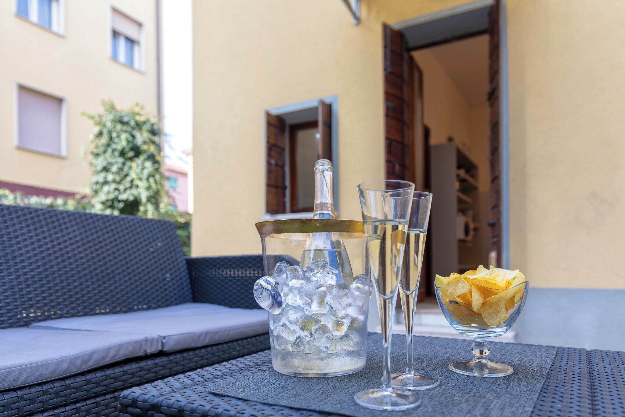 A clear glass pitcher filled with ice cubes, two glasses of a clear liquid, and a bowl of chips on a table outside.