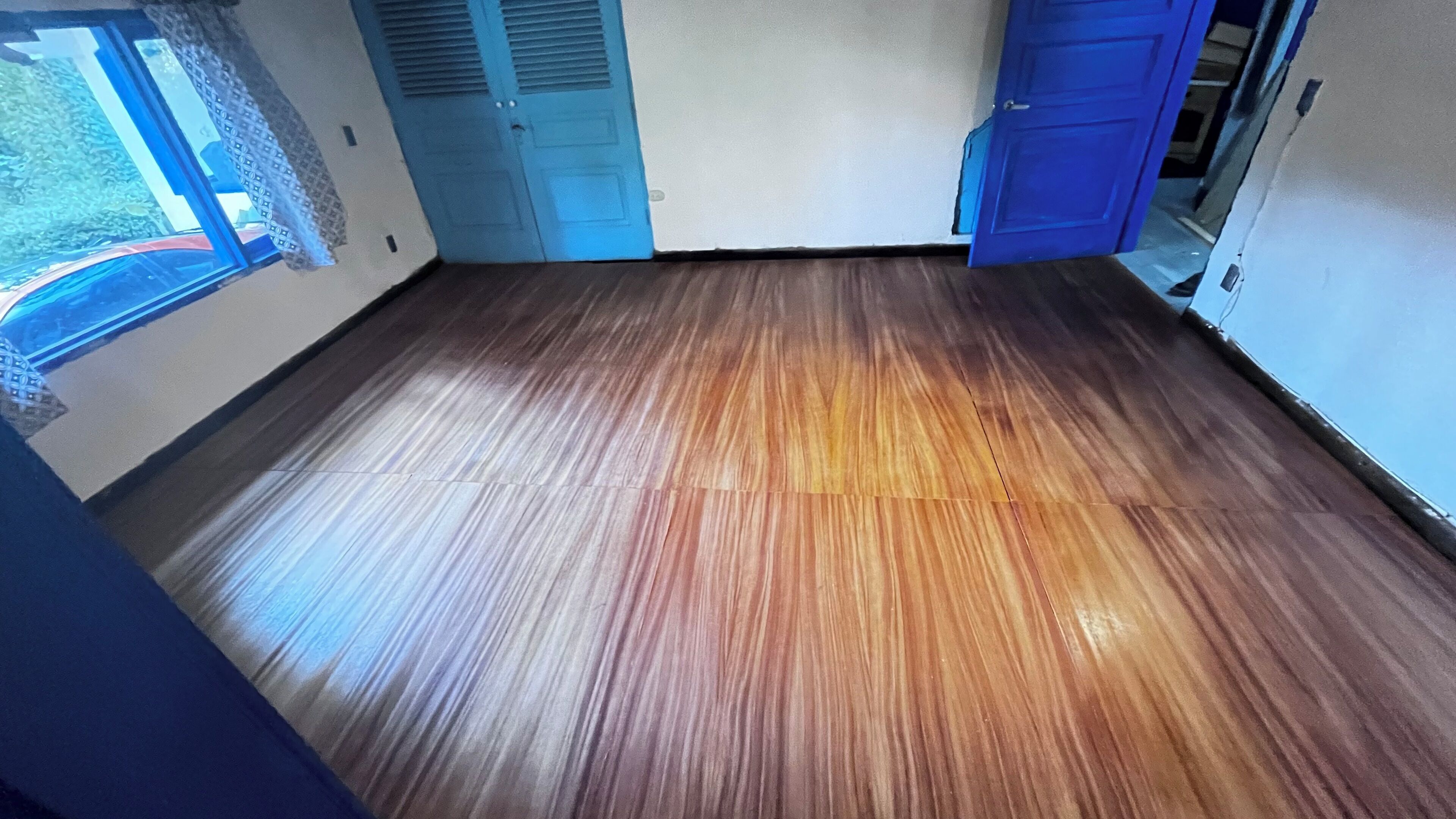 Una habitació amb terra de fusta, una porta blava i una finestra amb vistes.