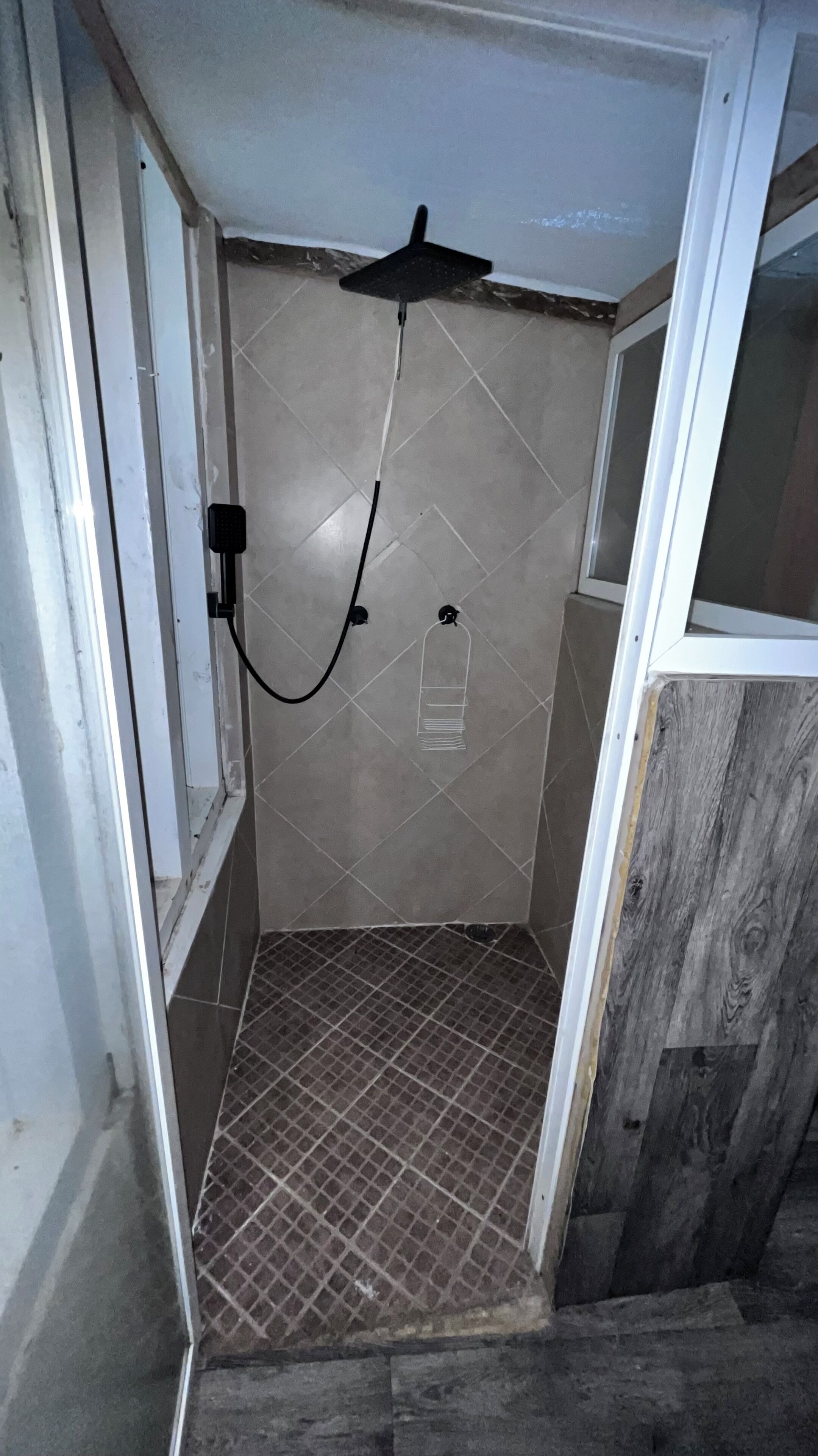 Ein kleiner Duschbereich mit Fliesen, einer Handbrause, einem sichtbaren Wasseranschluss und einem gefliesten Boden.