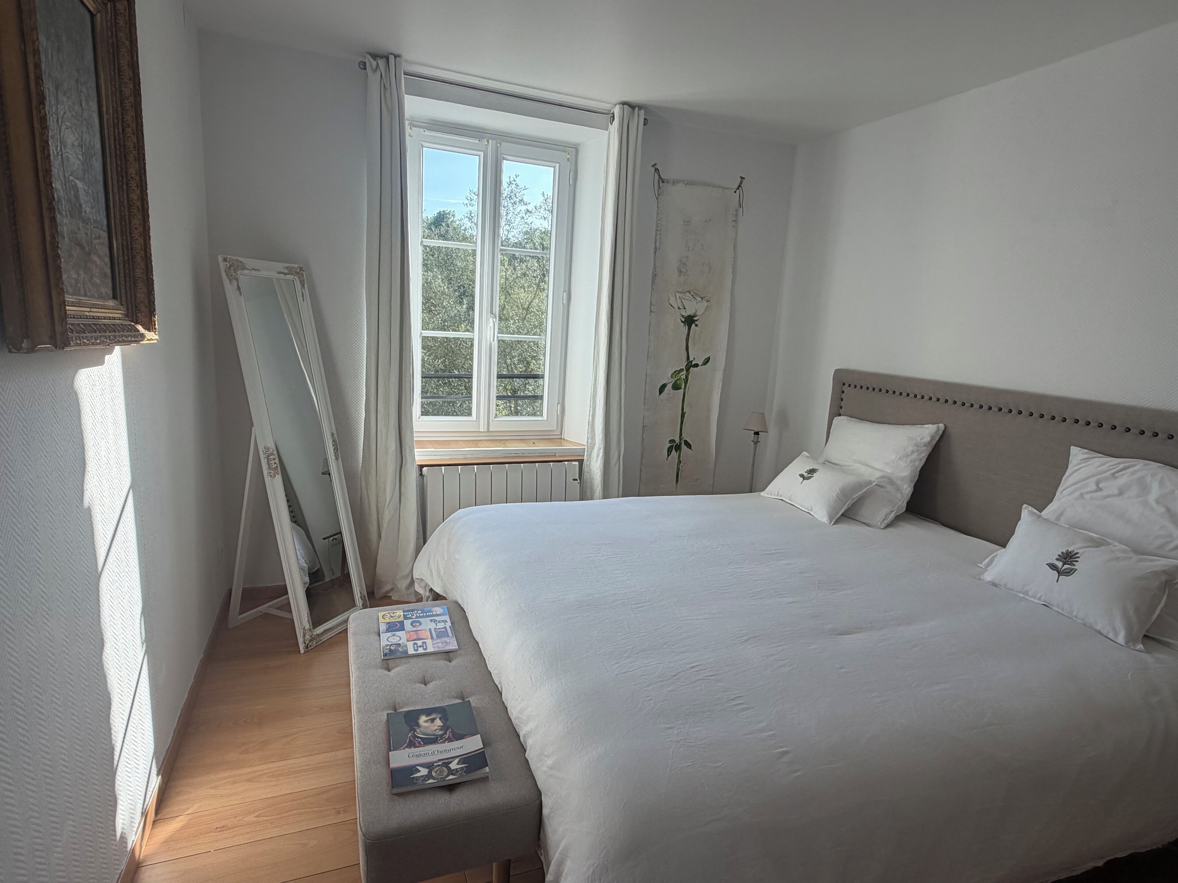 Een slaapkamer met een groot bed, een nachtkastje met boeken, een raam met gordijnen en een radiator.