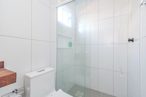 A modern bathroom with white tiles, a glass shower enclosure, a wooden vanity, and a toilet. - Apto Perto da Praia Com Piscina e Churrasqueira (Penha)