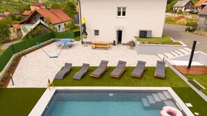 Ein Pool mit Liegestühlen und einem Tisch, umgeben von einem gepflegten Rasen und Blick auf eine Wohngegend.