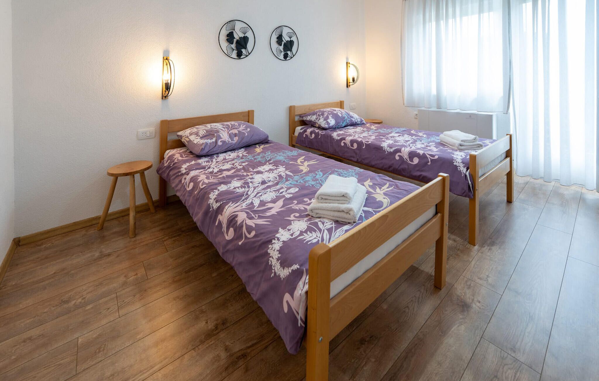 A room with two wooden beds, each with a purple floral bedspread and matching pillows, a small wooden stool, and wall-mounted light fixtures.