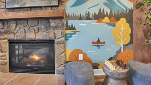 Chimenea encendida, pared de piedra, un mural con paisaje de lago y dos sillones.