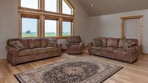 A spacious living room with a high ceiling, large windows, and a view of the countryside. There are three brown leather sofas and a matching armchair. - Teton View Retreat- Hot Tub - Game Room - Fire Pit (Alta)