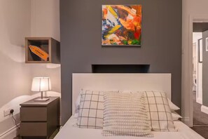 A bedroom with a bed, a nightstand, a lamp, a framed abstract painting, and a small shelf with a London flag. - Cosy 1-bedroom apartment in Newport perfect for relaxing stays (Newport)