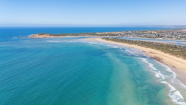 Vista aérea de una zona costera con playa, aguas azules cristalinas y un área residencial con casas.