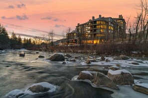 Een meerlaags hotel met een modern architectonisch ontwerp, gelegen naast een rivier met zichtbare rotsen en sneeuwplekken.