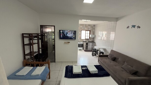 A living room with a sofa, a TV mounted on the wall, and a kitchen area in the background. - Angra Sol e Mar Suite: rest, comfort and enchanting beaches. Close to Brasfels. (Angra dos Reis)