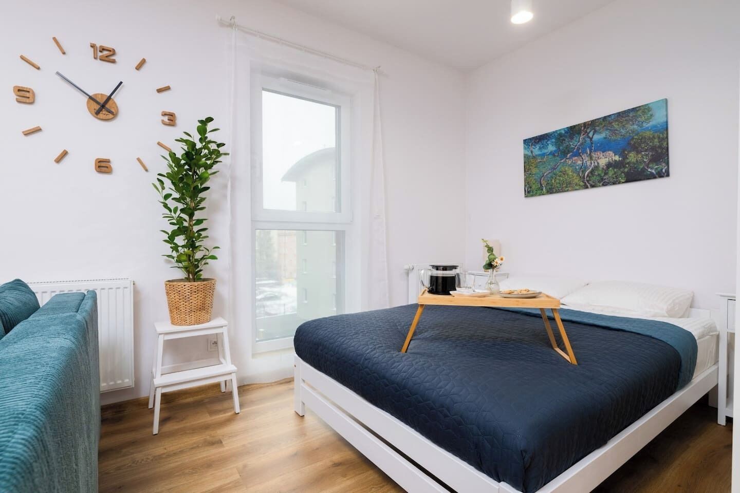 A modern bedroom with a bed, a small table with a coffee pot, a clock, a plant, and a painting on the wall.