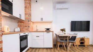 Una cocina compacta con armarios blancos, un muro de ladrillo decorativo, un horno empotrado y un comedor con mesa y sillas de madera.