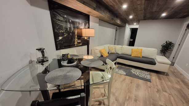 A modern living room with a glass dining table, a sofa with decorative pillows, a cityscape painting, and a wooden ceiling.