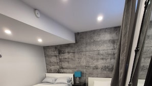 A bedroom with two beds, a nightstand with a lamp, a concrete accent wall, wooden flooring, and a woven basket with towels.