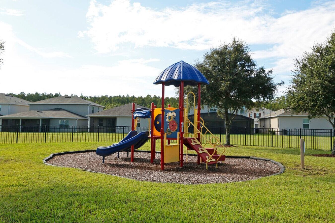 A playground with slides and climbing structures, surrounded by a fenced area and houses in the background.