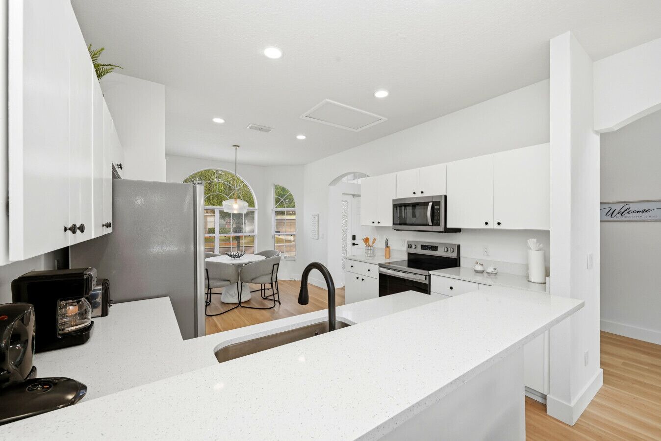 A modern kitchen with white cabinetry, a central island, a dining area with a table and chairs, and a view of the outdoors through large windows.