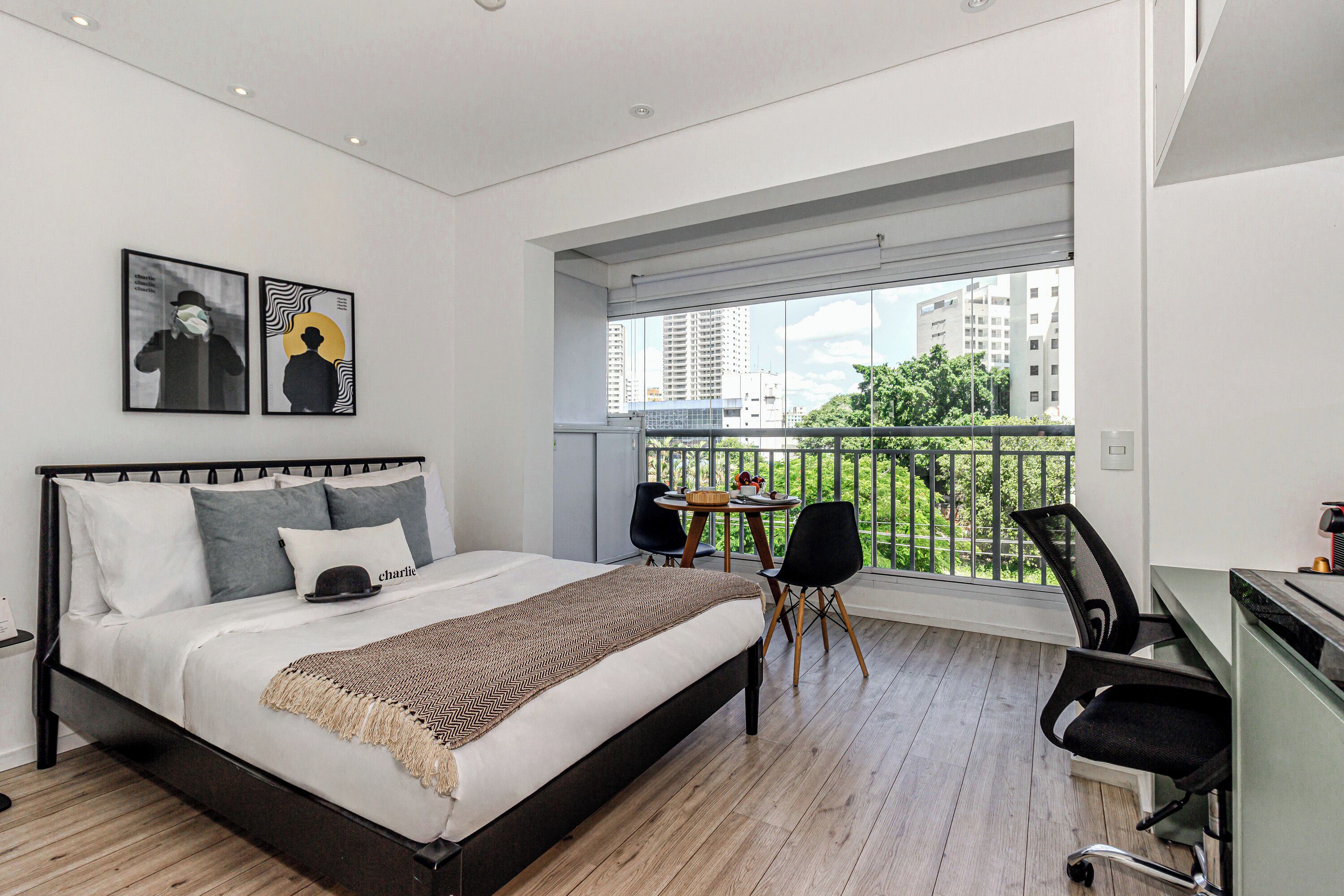 Un dormitorio moderno con una cama grande, un escritorio y un balcón con vistas a edificios y vegetación.