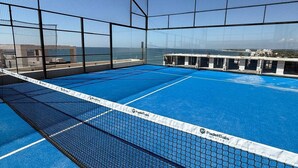 Ein Hallen-Tennisplatz mit Netz, umgeben von einem Maschendrahtzaun, mit Blick auf eine Küstenlandschaft.