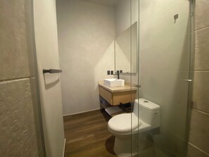 Un baño pequeño y moderno con suelo de madera, inodoro blanco, cabina de ducha de vidrio y tocador de madera con lavamanos blanco.