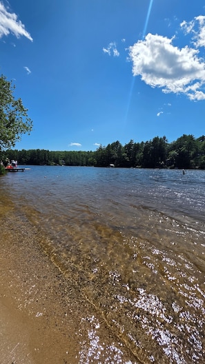 Ein klarer See mit sandigem Ufer, umgeben von Bäumen und blauem Himmel.