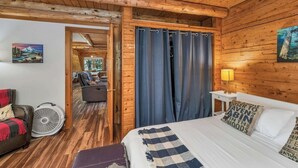 A wooden cabin room with a bed, a fan, a chair, and a view of a forest through a window. - Charming year round lake view cabin! (Wakefield)