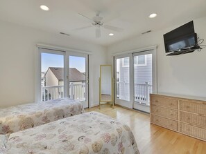 Quarto com duas camas, ventilador de teto, televisão e janelas amplas.