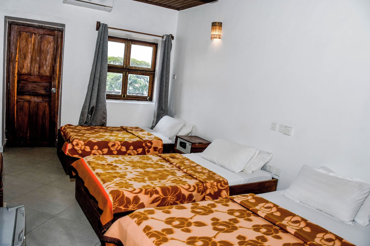 Une chambre d’hôtel avec trois lits, une fenêtre avec des rideaux, un luminaire fixé au mur et une porte en bois.